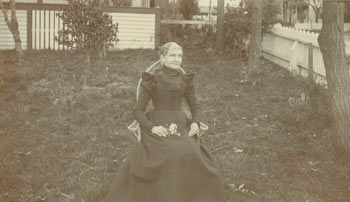 Item #63-2754 Monochrome Photograph of a woman seated in chair in backyard formally dressed (mourning wear?), holding flowers in her lap. 20th Century American Photographer.